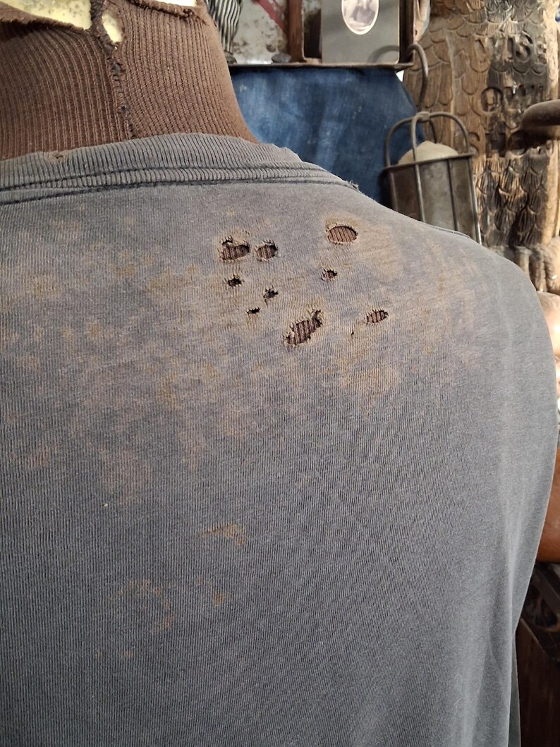 Puede incluir: Una camiseta azul descolorida con varios agujeros en la parte superior de la espalda. La camiseta parece desgastada y tiene una est&eacute;tica vintage. La tela muestra signos de desgaste, con decoloraci&oacute;n y manchas.