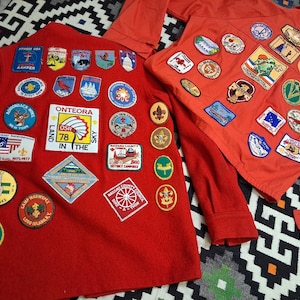 May include: Two red long-sleeved shirts adorned with numerous patches. The patches display diverse designs, including badges with text, flags, and illustrations of animals and landscapes. The shirts are likely associated with scouting or a similar youth organisation.