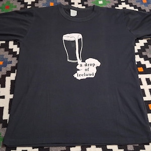 Op de afbeelding: Donkergrijs t-shirt met een witte afbeelding van een pint bier en de omtrek van Ierland, met de tekst "a drop of Ireland" eronder. Het shirt ligt plat op een patroon oppervlak.
