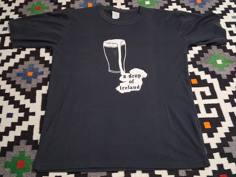 Op de afbeelding: Donkergrijs t-shirt met een witte afbeelding van een pint bier en de omtrek van Ierland, met de tekst "a drop of Ireland" eronder. Het shirt ligt plat op een patroon oppervlak.