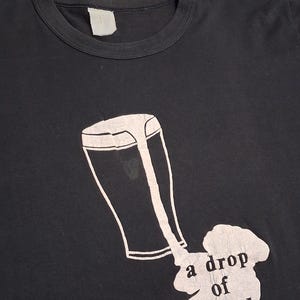 Op de afbeelding: Zwart T-shirt met een witte afbeelding van een pint bier die over een kaart van Ierland wordt gemorst. De tekst "a drop of Ireland" staat eronder. Het shirt heeft een ronde hals en een blanco label.