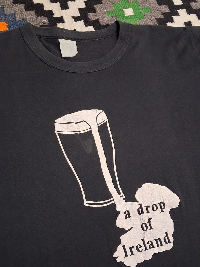 Op de afbeelding: Zwart T-shirt met een witte afbeelding van een pint bier die over een kaart van Ierland wordt gemorst. De tekst "a drop of Ireland" staat eronder. Het shirt heeft een ronde hals en een blanco label.