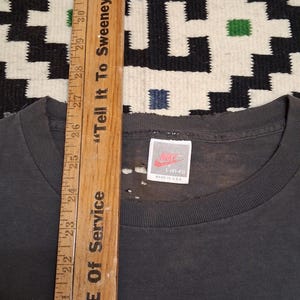 Puede incluir: Una camiseta gris oscuro con cuello redondo y una etiqueta Nike. La camiseta es talla L (41-43) y est&aacute; hecha en los EE. UU. Una regla de madera se coloca verticalmente junto a la camiseta, con el texto "Tell It To Sweeney" y "Measure Of Service".