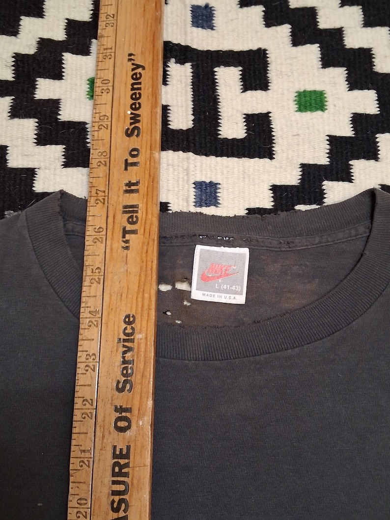 Puede incluir: Una camiseta gris oscuro con cuello redondo y una etiqueta Nike. La camiseta es talla L (41-43) y est&aacute; hecha en los EE. UU. Una regla de madera se coloca verticalmente junto a la camiseta, con el texto "Tell It To Sweeney" y "Measure Of Service".