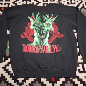 Può includere: Felpa nera con grafica di un demone verde e dettagli rossi, con la scritta "ROOT OF ALL EVIL". Il disegno mostra una faccia di demone che digrigna i denti con corna e occhi rossi. La felpa è probabilmente realizzata in un materiale morbido e confortevole.