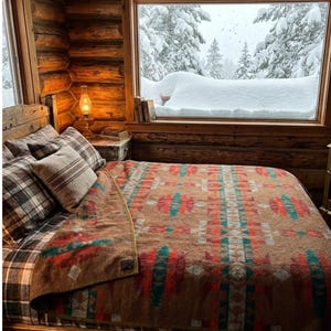Könnte beinhalten: Ein Bett mit einer braun, rot und blaugrün gemusterten Decke, karierten Kissen und einem beigen Kissen. Das Bett steht in einem Blockhaus mit einem Fenster, das eine verschneite Waldlandschaft zeigt, einer kleinen Lampe und Büchern auf einem Nachttisch.