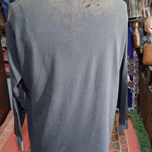 Puede incluir: Una camiseta de manga larga azul gris&aacute;ceo descolorido con detalles desgastados en la parte superior de la espalda. Las mangas tienen un pu&ntilde;o azul en contraste. La camiseta se muestra en un maniqu&iacute;.