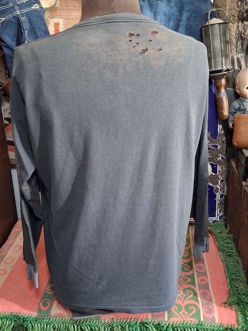 Puede incluir: Una camiseta de manga larga azul gris&aacute;ceo descolorido con detalles desgastados en la parte superior de la espalda. Las mangas tienen un pu&ntilde;o azul en contraste. La camiseta se muestra en un maniqu&iacute;.