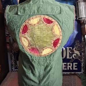 May include: An olive green sleeveless jacket featuring a large circular patch on the back. The patch consists of a central olive green velvet circle surrounded by alternating yellow and deep red velvet petal shapes with gold embroidery trim.