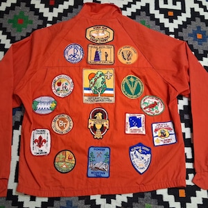 May include: A bright orange jacket adorned with numerous embroidered patches. The patches display diverse designs, including a tree, a Native American head, and a mountain landscape. The jacket features a zip closure and long sleeves. The patches include text such as "1973 National Scout Jamboree".