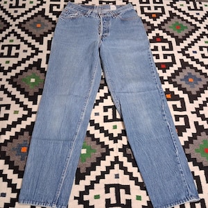 May include: Light blue denim jeans featuring a high-waisted fit, a straight leg silhouette, and a button-fly closure. The jeans have a classic five-pocket design with visible stitching along the seams and a faded wash finish throughout the fabric.