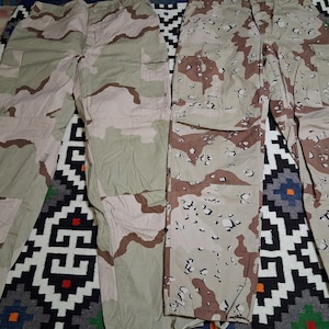 May include: Two pairs of camouflage trousers in desert tan, brown, and green shades. The trousers have multiple pockets and a button and zip closure. The fabric displays a military-style pattern.