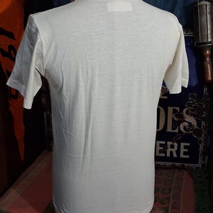 May include: Cream-colored, short-sleeved t-shirt displayed on a mannequin. The shirt has a simple, classic design with a crew neck and a rectangular tag at the back of the neck. The fabric appears to be lightweight and slightly textured.