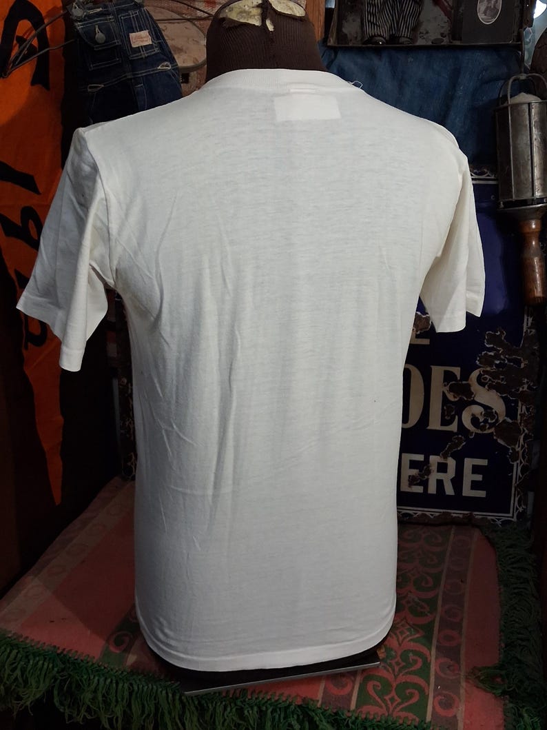 May include: Cream-colored, short-sleeved t-shirt displayed on a mannequin. The shirt has a simple, classic design with a crew neck and a rectangular tag at the back of the neck. The fabric appears to be lightweight and slightly textured.