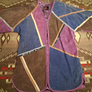 May include: A patchwork suede jacket featuring an open front and short sleeves. The garment is constructed from angular panels of brown, purple, and blue suede leather, accented with thin, metallic-textured piping along the seams and a purple trim.