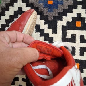 May include: Red and white Nike sneakers with white laces. The sneakers have a red upper with a white swoosh and the Nike logo. The sole is off-white. The sneakers are on a patterned rug.