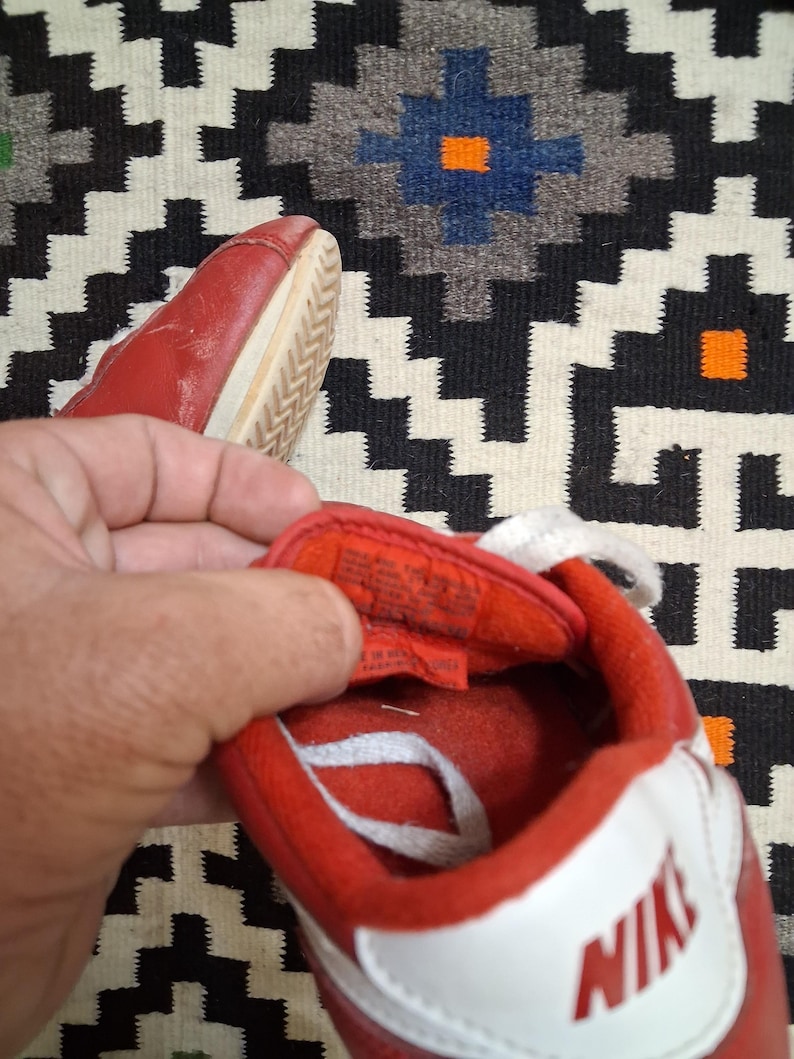May include: Red and white Nike sneakers with white laces. The sneakers have a red upper with a white swoosh and the Nike logo. The sole is off-white. The sneakers are on a patterned rug.