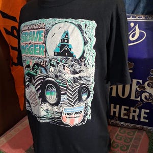 May include: Black t-shirt featuring a graphic of a monster truck with the text "Dennis Anderson's Grave Digger". The design includes a haunted house, a full moon, and the "United States Hot Rod Association" logo.