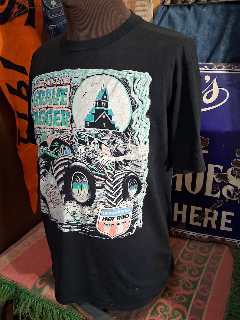May include: Black t-shirt featuring a graphic of a monster truck with the text "Dennis Anderson's Grave Digger". The design includes a haunted house, a full moon, and the "United States Hot Rod Association" logo.