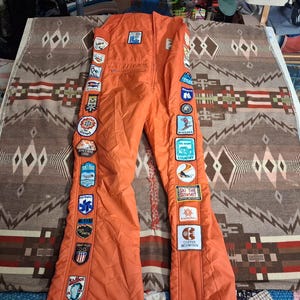 Puede incluir: Un traje de esquí de una pieza de color naranja brillante con una fila vertical de parches bordados coloridos a lo largo del lado exterior de cada pierna. Los parches muestran varios logotipos de estaciones de esquí y el texto Ski The Summit.