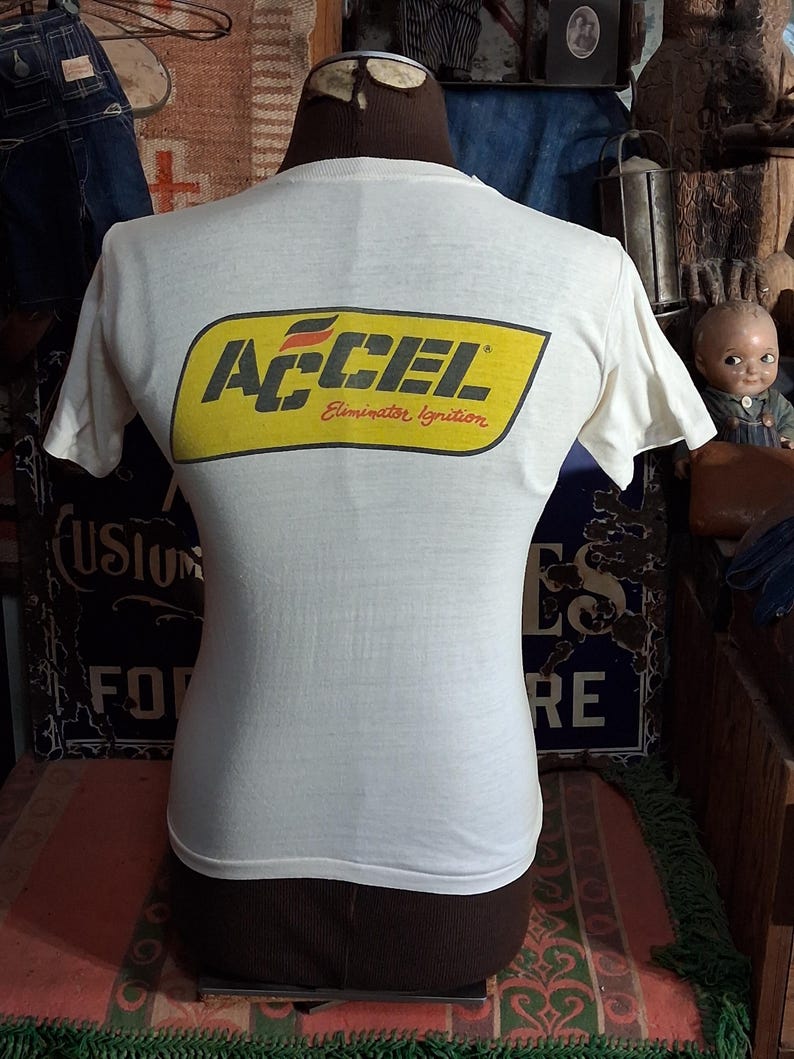 K&ouml;nnte beinhalten: Wei&szlig;es T-Shirt mit gelb-schwarzem ACCEL Eliminator Ignition Logo auf dem R&uuml;cken. Das T-Shirt wird auf einer Schaufensterpuppe pr&auml;sentiert. Das Vintage-T-Shirt hat kurze &Auml;rmel und einen Rundhalsausschnitt. Das Design ist im Retro-Stil.