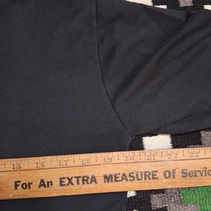 Op de afbeelding: Close-up van een zwarte t-shirtmouw met een houten liniaal over de onderkant. De liniaal heeft de tekst "For An EXTRA MEASURE Of Service". De liniaal meet van 38 tot 58 cm.