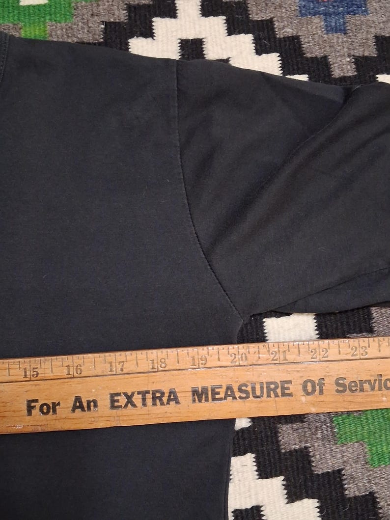 Op de afbeelding: Close-up van een zwarte t-shirtmouw met een houten liniaal over de onderkant. De liniaal heeft de tekst "For An EXTRA MEASURE Of Service". De liniaal meet van 38 tot 58 cm.