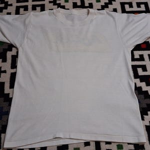 K&ouml;nnte beinhalten: Ein schlichtes wei&szlig;es T-Shirt mit kurzen &Auml;rmeln, flach hingelegt. Das Shirt besteht aus einem weichen, wahrscheinlich Baumwollmaterial. Der Kragen ist ein einfacher Rundhalsausschnitt. Das Shirt ist sauber und scheint ungetragen zu sein.