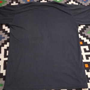 Op de afbeelding: Een donkergrijs T-shirt met korte mouwen. Het shirt heeft een ronde hals en is gemaakt van een zacht materiaal. Het T-shirt wordt gepresenteerd op een patroonkleed met zwart, wit en andere kleuren.