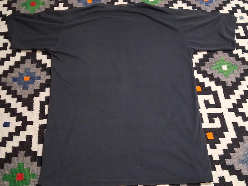 Op de afbeelding: Een donkergrijs T-shirt met korte mouwen. Het shirt heeft een ronde hals en is gemaakt van een zacht materiaal. Het T-shirt wordt gepresenteerd op een patroonkleed met zwart, wit en andere kleuren.