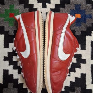 May include: Pair of red leather sneakers with white accents, including a large white swoosh on the side. The shoes have white laces and a cream-colored sole with a red stripe. The sneakers are displayed on a patterned rug.