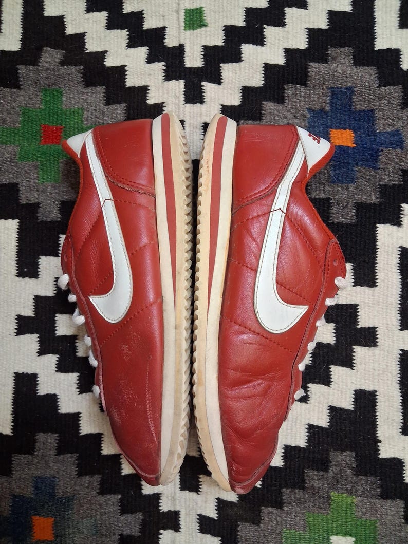 May include: Pair of red leather sneakers with white accents, including a large white swoosh on the side. The shoes have white laces and a cream-colored sole with a red stripe. The sneakers are displayed on a patterned rug.