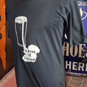 Op de afbeelding: Donkergrijs t-shirt met een witte afbeelding van een pint bier en de tekst "a drop of Ireland". Het shirt heeft een ronde hals en korte mouwen. Het shirt wordt op een mannequin getoond.