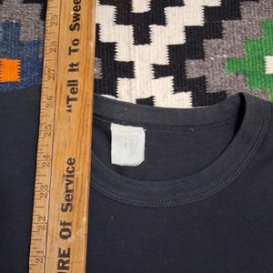 Op de afbeelding: Een zwart T-shirt met een ronde hals. Een houten liniaal met de tekst "Tell It To Sweeney" en "EASURE Of Service" ligt naast het shirt. De achtergrond is een patroonkleed.