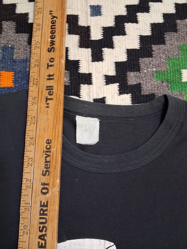 Op de afbeelding: Een zwart T-shirt met een ronde hals. Een houten liniaal met de tekst "Tell It To Sweeney" en "EASURE Of Service" ligt naast het shirt. De achtergrond is een patroonkleed.