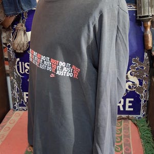 Puede incluir: Una camiseta gris de manga larga con el logo de Nike y la frase "JUST DO IT" en rojo y blanco. La camiseta se exhibe en un maniqu&iacute;. Tiene cuello redondo y mangas largas.