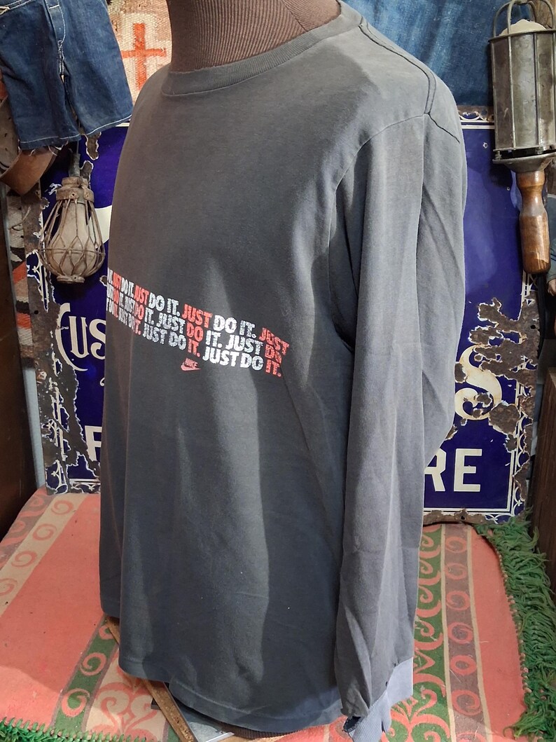 Puede incluir: Una camiseta gris de manga larga con el logo de Nike y la frase "JUST DO IT" en rojo y blanco. La camiseta se exhibe en un maniqu&iacute;. Tiene cuello redondo y mangas largas.