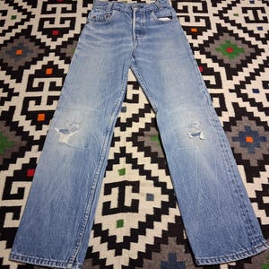 Può includere: Jeans in denim blu chiaro con chiusura a bottoni e dettagli strappati. I jeans hanno una vestibilità ampia e presentano toppe strappate sulle ginocchia. I jeans sono stesi su un tappeto fantasia.