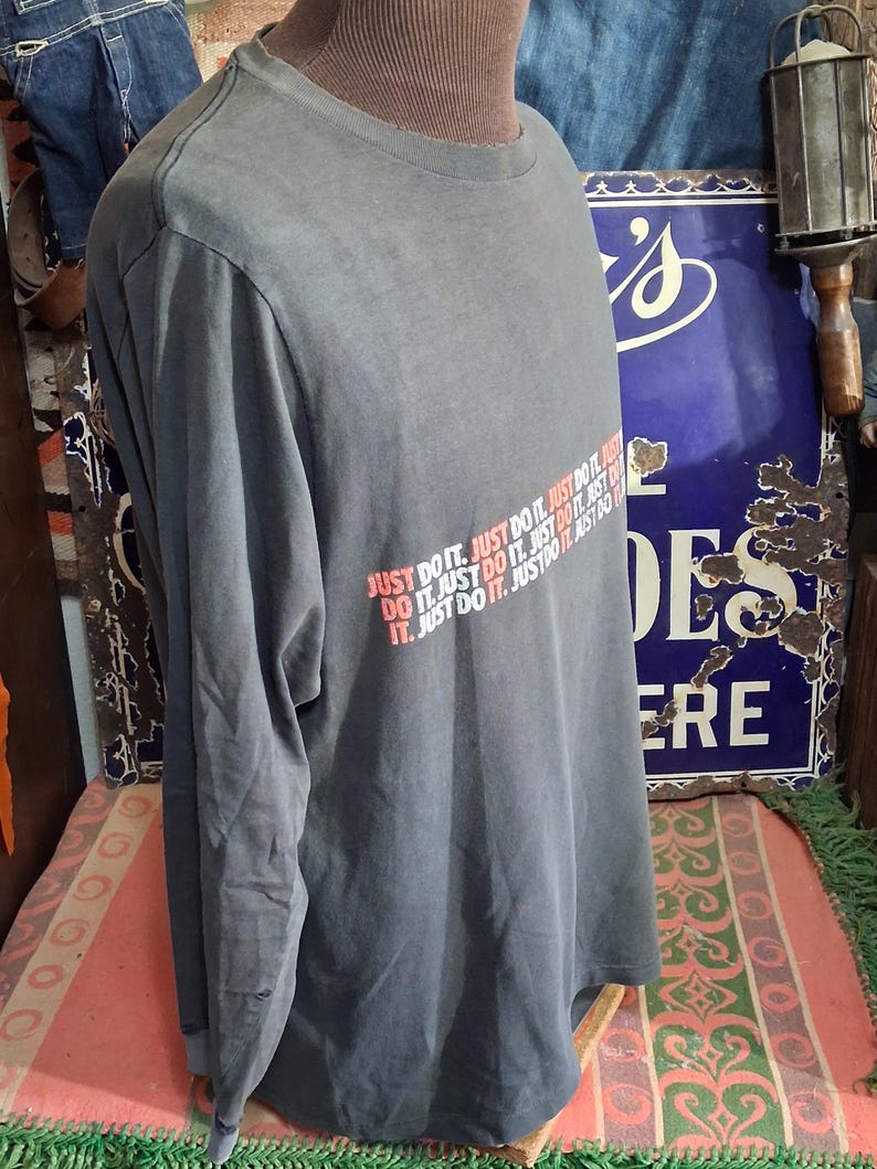 Puede incluir: Una camiseta gris de manga larga con el texto "JUST DO IT" en rojo y blanco. La camiseta se muestra en un maniqu&iacute;. La camiseta es de estilo casual.