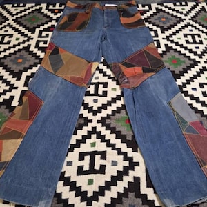 May include: Blue denim jeans with patchwork leather detailing in brown, tan, and burgundy. The jeans have a classic five-pocket design and a flared leg. The patchwork is on the front and back of the jeans.