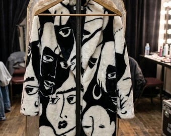 Peut inclure: Un manteau à manches longues avec un imprimé graphique noir et blanc représentant des visages humains stylisés. Le manteau est sur un cintre en bois avec une étiquette indiquant : ARTIST: JOSHUA - BACKSTAGE COSTUME. Il possède un col à revers.