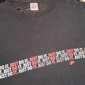 Puede incluir: Camiseta gris oscuro con el logo de Nike y la frase "JUST DO IT" impresa en blanco y rojo en diagonal. La camiseta tiene un aspecto desgastado y vintage.