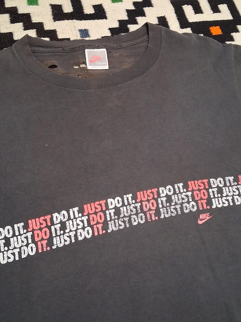 Puede incluir: Camiseta gris oscuro con el logo de Nike y la frase "JUST DO IT" impresa en blanco y rojo en diagonal. La camiseta tiene un aspecto desgastado y vintage.