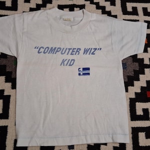 Op de afbeelding: Een wit T-shirt met korte mouwen en de blauwe tekst "COMPUTER WIZ" KID op de borst gedrukt. Onder de tekst staat een klein blauw vierkant logo met de witte tekst "ONE UP" erin. Het T-shirt heeft een ronde hals en een standaard zoom.