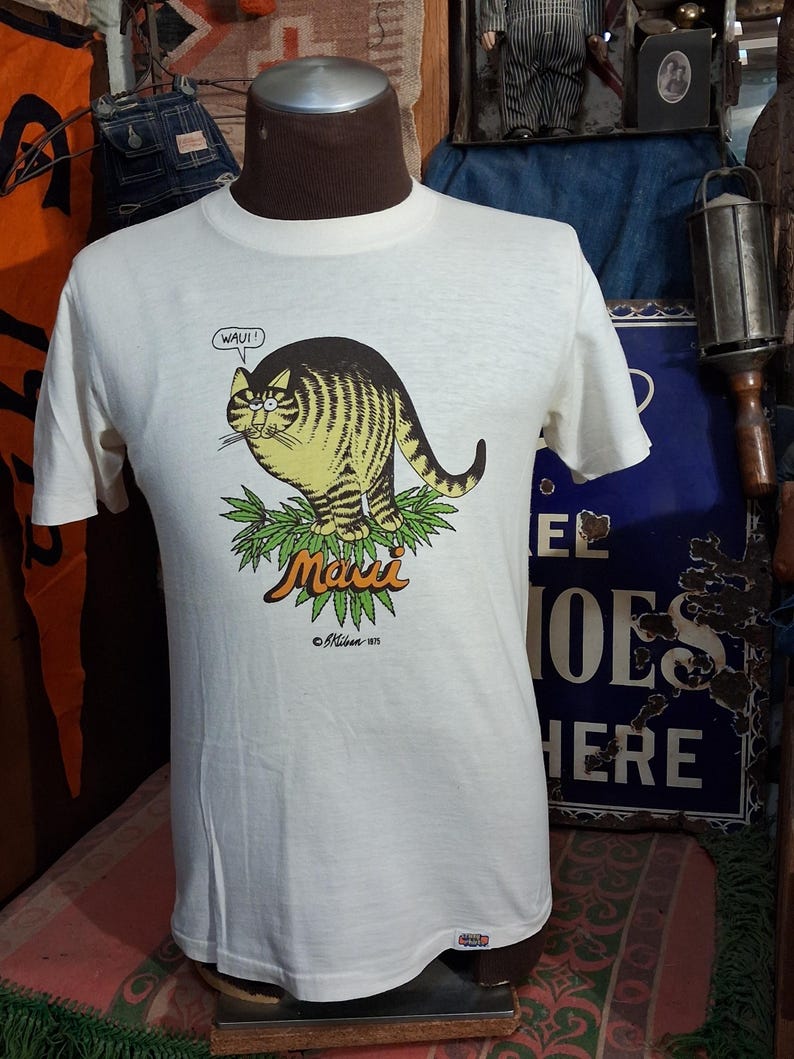 May include: Off-white t-shirt featuring a cartoon cat with yellow and brown stripes, sitting on green leaves. The text "Maui" is above the leaves, and "WAUI!" is in a speech bubble. The shirt is from &copy;Mibon 1975.