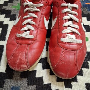 May include: Pair of red leather sneakers with white laces and a white Nike swoosh logo on the sides. The shoes have a worn appearance, with some scuff marks on the toe area. The soles are off-white.