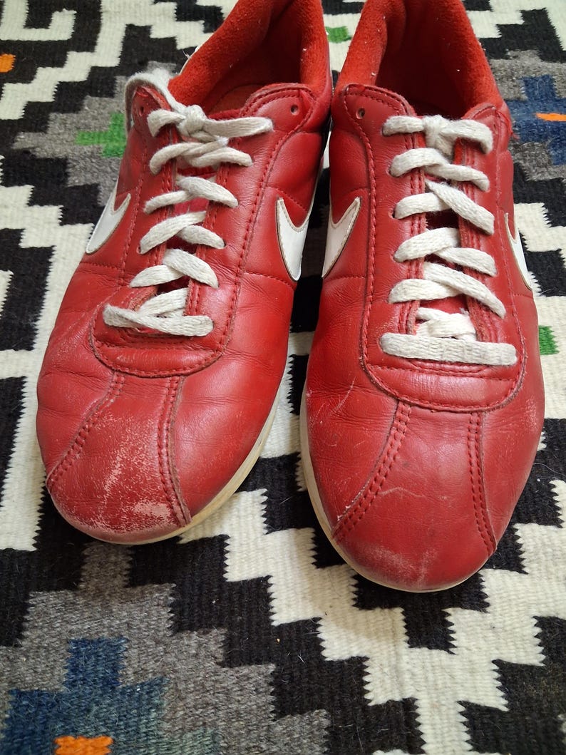 May include: Pair of red leather sneakers with white laces and a white Nike swoosh logo on the sides. The shoes have a worn appearance, with some scuff marks on the toe area. The soles are off-white.