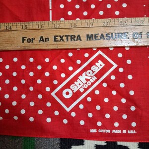 K&ouml;nnte beinhalten: Rotes und wei&szlig; gepunktetes Bandana mit dem Text "OSHKOSH B'GOSH" in einem wei&szlig;en Rechteck. Das Bandana hat auch den Text "100% Cotton Made in U.S.A." und "K WEAR". Ein Holzlineal liegt oben auf.
