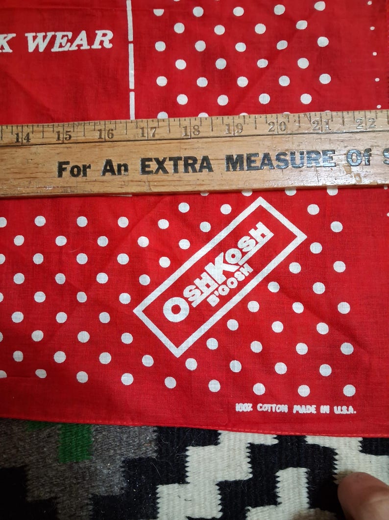 K&ouml;nnte beinhalten: Rotes und wei&szlig; gepunktetes Bandana mit dem Text "OSHKOSH B'GOSH" in einem wei&szlig;en Rechteck. Das Bandana hat auch den Text "100% Cotton Made in U.S.A." und "K WEAR". Ein Holzlineal liegt oben auf.