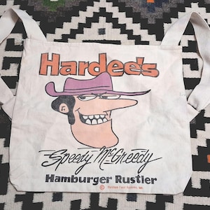 Può includere: Borsa tote in tela bianco sporco con il logo Hardee's e Speedy McGreedy, un personaggio cowboy dei cartoni animati. La borsa ha lunghe cinghie e la scritta "Hamburger Rustler" sotto.
